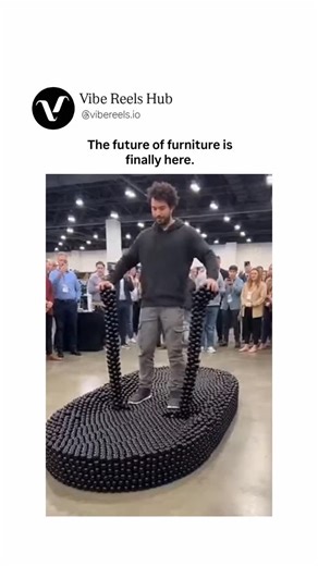 Vibe Reels Hub™ on Instagram: "The video showcases a groundbreaking demonstration of adaptive, shape-shifting furniture at a large indoor convention or expo hall, with bright overhead lights and a massive crowd of onlookers in business casual attire gathered around. A bearded man in a dark sweater and gray pants stands confidently atop an enormous, black, modular structure composed of countless small interlocking hexagonal or conical elements, forming a fluid, organic base that resembles a high-
