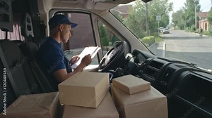 Delivery man in uniform sitting in car, taking package and making marks before delivering it to a client