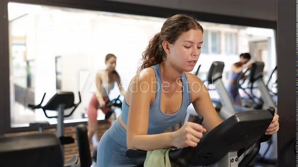 Positive young woman doing spin bike exercises in well-equipped gym during training session