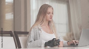 A young woman is using a robotic prosthetic arm hand attached to her body to work on a desk. she is doing computer document and communication work. woman tries to control the smart arm in her home.