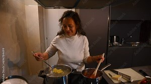 Charming female chef, housewife preparing delicious pasta for dinner at home kitchen. Happy woman mixing farfalle in boiling water and stirring tomato sauce in a saucepan, standing by electric stove