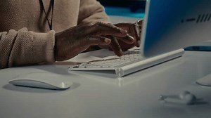 Close-up of hands of anonymous middle-aged African American male IT specialist in beige sweater, with badge typing on computer keyboard and using mouse while working in office