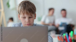 Schoolboy using laptop and sitting at table while learning online at home, parents are behind spbi. Close-up view of boy looks at screen of device and studies remotely, sits at desk in light room, dad