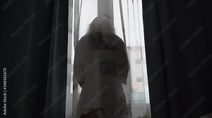 Silhouette of woman against window behind transparent curtain, woman in dressing gown looking out the window and using her smart phone. Stock Video