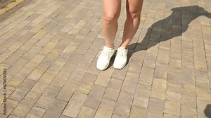 View of a woman's legs in white sneakers walking along the paving stones in a park in summer. slow motion woman walking