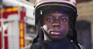 Portrait of African American firefighter approaches from fire van with flashing lights on and standing in front of camera looking. Concept of saving lives, heroic profession, fire safety