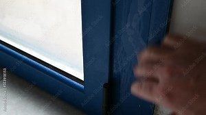 Thermal insulation problems cause condensation on the door window frame: the Caucasian man's hand passes over the blue metal frame, highlighting the formation of humidity.