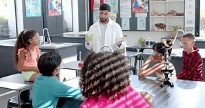 In a school classroom, a biracial teacher demonstrates a chemistry experiment. Surrounded by diverse students, he holds a beaker, while kids engage with microscopes and observe, unaltered.