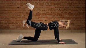 Executing precise donkey kick pulses with her right leg, the professional fitness trainer focuses on glute toning and building endurance, against the structured elegance of a brick-walled gym.