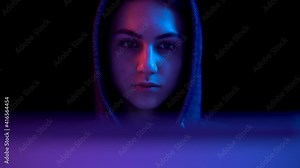 Young woman in a hood with a laptop. Hacker makes a hack through a laptop. Blue and red light falls on a woman on a black background. The camera goes down.