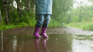 Little child jumps in puddle on park road closeup. Funny little boy plays splashing water water on rainy spring day. Outdoors activity and happy childhood
