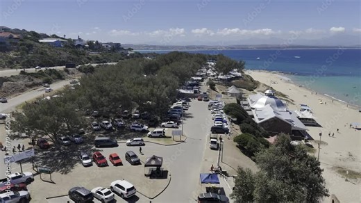 Drone flies north and follows Santos Beach over parking lot and fully packed campervan area, then flies out to the sea on a sunny day on the Garden Route in Mossel Bay, South Africa