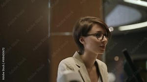 Pretty female receptionist in eyeglasses working at front desk in hotel and talking to guests during check-in procedure