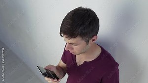 Camera shoots from the top upset young man looking at smartphone screen, dissatisfied with bad news message, spam or scam sms. Unhappy stressed male mobile app user getting phishing link in email or l