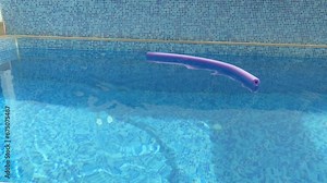 A clip of a blue mosaic tile swimming pool. A purple pool noodle is thrown in the water making many ripples in the water. The noodle stays floating in the water.