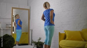 Lovely woman in casual clothes checking her body in mirror, feeling excited and beautiful after successful weight loss program in domestic room. Joyful female admiring her reflection in mirror.