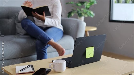 Laptop, notebook, pen, phone and cup on the table with a woman relaxing on the sofa reading a book in the background