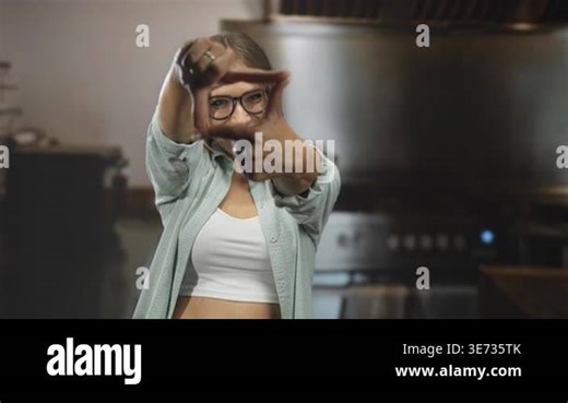 Woman framing face with hands and glasses in a kitchen building, smiling while wearing open shirt and white crop top; playful focus Stock Video Footage - Alamy