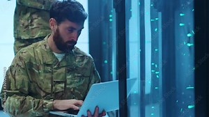 Bearded military man opens a server cabinet and downloading secret information into laptop. In background armed officer with gun guarding and cooperating in data center.