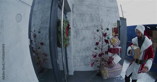 Santa Claus Delivers Package to Woman at Doorstep. Male Courier Uses Payment Terminal For Contactless Transaction, Captured By Doorbell Camera. Festive Blue Van Full of Gifts Visible Behind Him.