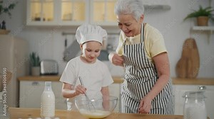 Senior grandma in apron touches shows her granddaughter how to roll out dough using rolling pin in kitchen at home. Family cooking together, homemade bakery, pastry, sweets concept. Girl in cooks cap.