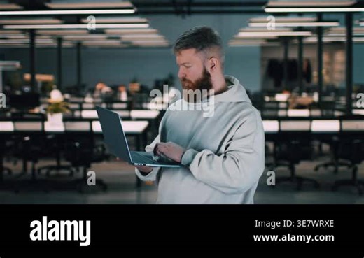 The man stands in an office environment using a laptop. The space features multiple desks with bright overhead lights and no people around Stock Video Footage - Alamy