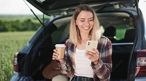 Front view of blonde woman sitting in car trunk, using smartphone, drinking coffee. Young female chatting, texting, messaging, holding paper cup. Concept of modern lifestyle.
