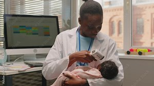 Medium shot of young African American female pediatrician holding little baby in arms and checking her temperature with infrared thermometer in modern doctors office