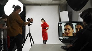Model and production team in the studio. Female posing for photographer, girl assistant fixes makeup and hair, editor at the desktop checking photos.