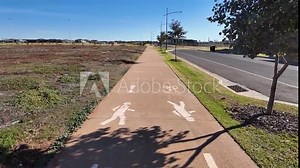 A shared-use path marked with pedestrian and bicycle symbols, alongside a new road in a recently developed suburban neighborhood with open undeveloped lots and young trees in Aintree, Australia.