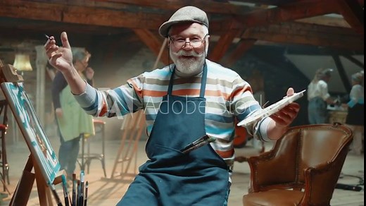Elderly artist man is sitting on a stool at studio in front of canvas, and working with inspiration, painting a picture, then looks at camera and smiles