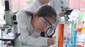 Young scientist girl use magnifying glass to look at chemical test tube and teacher also tell them to analysis the reaction in laboratory or classroom with glass windows and happy education concept.