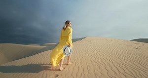 Cinematic slow motion of woman walking by sand dune. Barefoot female traveler in waving on the wind dress by the rippled sand desert surface with dark grey cloud on background. 4K Scenic nature