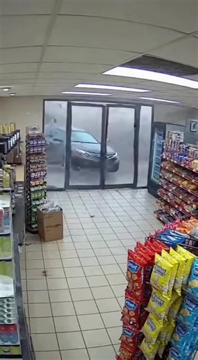 CCTV footage shows the inside of a small convenience store from a fixed ceiling camera as a powerful tornado moves directly through the area. In the first moments, the lights flicker and the entire building begins to shake. The front windows suddenly implode, sending glass and debris flying across the floor. Shelves rattle violently as bags of chips, candy, and boxed items are ripped free and hurled through the air. Coolers burst open and bottled drinks explode across the aisle, liquid spraying 