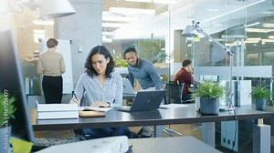 Busy International Office, Beautiful Hispanic Woman Working at Her Desk on a Laptop, She Waves Hello to the Passing Colleague, in the Background Her Coworkers Have Work Related Conversations.