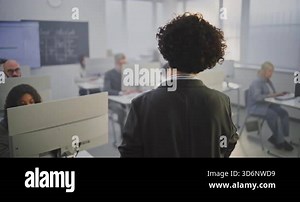 Female Teacher in Elegant Attire Guides Adult Learners During Programming Class. Classroom Filled With Concentrated Students Working on Computers. Empowerment Through Digital Skills. Back View.