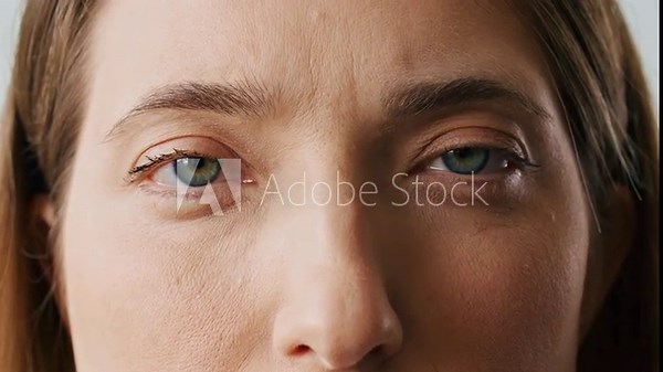 Very close camera view of Caucasian girls face. Woman with charming green eyes and straight brown hair. Not expressing any emotions. Wearing makeup. Looking directly at camera. Blinking eyes.