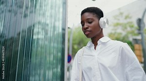 young charming Black woman in headphones, checks her smartwatch device with surprised expression as she rushes to work. Technology, modern city life and hustle of professional responsibilities