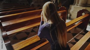 This video shows a young caucasian woman kneeling and praying at beautiful catholic church pew, rear view with sun beaming down.