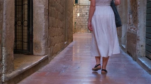 Woman exploring narrow old town alley at night. Unrecognizable woman wearing a summer dress walking away from the camera through a narrow, illuminated stone alley in a historic european old town
