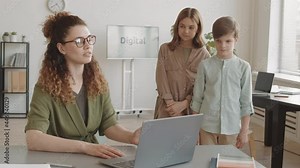 Medium long of two guilty Caucasian boy and girl standing by desk of young angry Caucasian teacher, lecturing students for bad homework