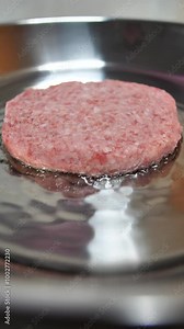 Vertical: Juicy large beef hamburger patty is fried in a stainless steel frying pan. Cooking a delicious burger with a meat patty. Fast food and a snack on a lunch break