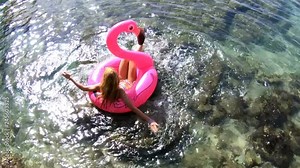 Young slender woman wearing rainbow bikini enjoying the sea and the sun have a fun splashing water lying on an inflatable pink Flamingo. Top view. Slow motion
