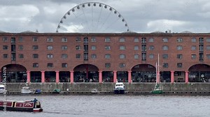 Boat passes the sight of the iconic Royal Albert Dock with the wheel of Liverpool towering above on cloudy overcast day - Liverpool, England, UK