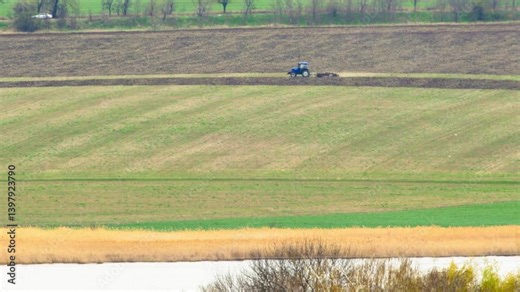 Spring agricultural work in Ukraine. Tractor plows agricultural land in countryside, preparing soil for planting. Wide rural landscape shows tractor working on farmland during early spring.