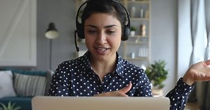 Happy indian young woman wear headset communicating by conference call speak looking at computer at home office, video chat job interview or distance language course class with online teacher concept