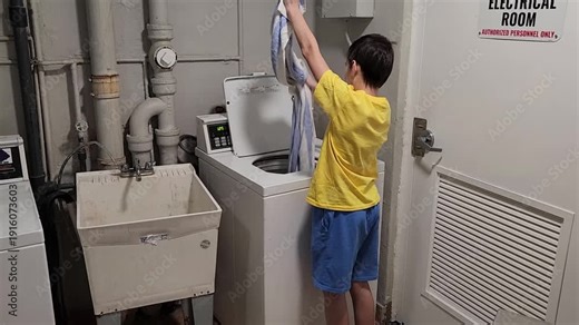 Young man takes clean wet laundry out of a washing machine and shakes it to loosen the fabric before drying. The action highlights routine laundry care, clothing handling, and everyday household work.