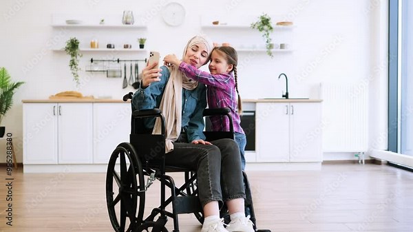 Bright, airy setting highlights family bonding, technology use, and inclusion. Muslim mother in wheelchair and daughter have video call, take selfie using phone in modern kitchen.