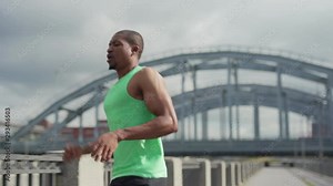 Young athletic African man in green tank top running on bridge during marathon training and stopping at railing to take break and catch breath