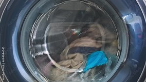 The washing machine washes dirty colored clothes. Washing clothes in a household washing machine. Close-up of the rotation of a drum washing machine.